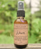 A 2 oz amber glass spray bottle of Soulgasm Weaver Smokeless Sacred Spray, resting on a weathered wooden surface with a soft-focus green garden background.