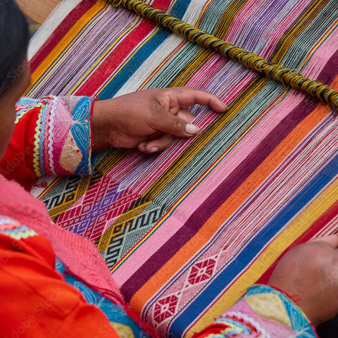 A close-up of a person weaving a brightly striped textile with pink, orange, yellow, blue, green, and red bands, wearing a colorful embroidered garment. The scene shows hands guiding the weaving process.