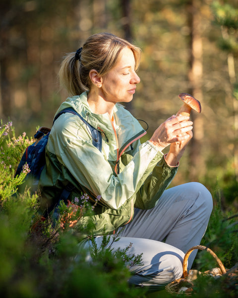 A woman in a green hooded jacket and gray pants sits in a sunlit forest clearing, holding a mushroom near her face. She has a dark blue backpack on her back, with trees and greenery blurred in the background.
