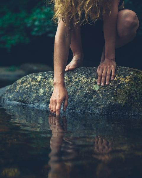 A woman crouches on a mossy rock in a dark forest stream, reaching her hand toward the water. Her long blonde hair is visible, and her bare feet and hands are shown against the dark water and foliage.