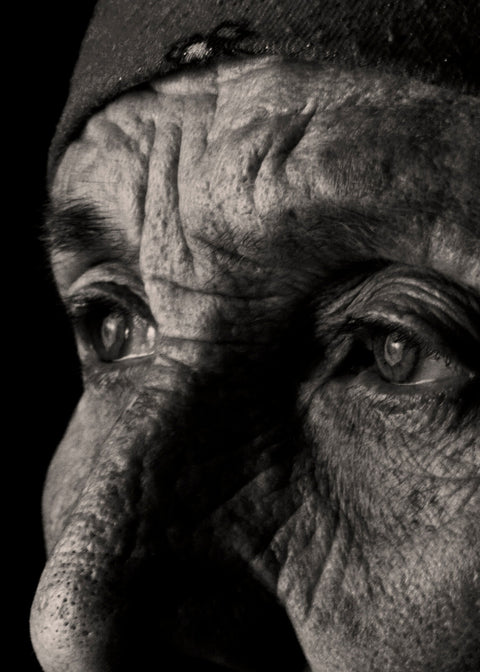 A dramatic black and white close-up portrait of an elderly person's face, showing deep wrinkles and expressive eyes against a solid black background.