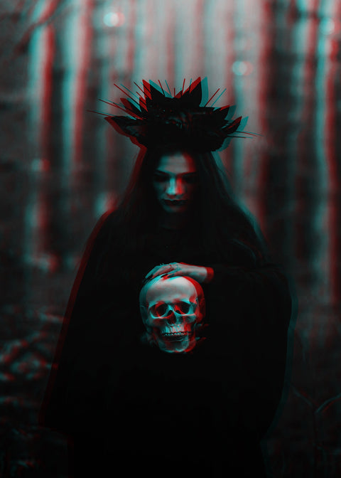 A dark, moody scene of a person in a black hooded robe standing in a forest, holding a skull in front of their face. The image has a strong red-cyan 3D anaglyph visual effect.