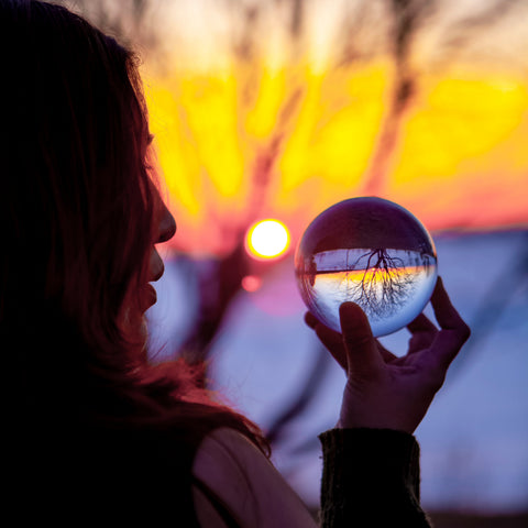 A woman in silhouette holds a clear glass crystal ball against a warm sunset sky, with tree branches and a glowing sun visible inside the sphere and in the background.