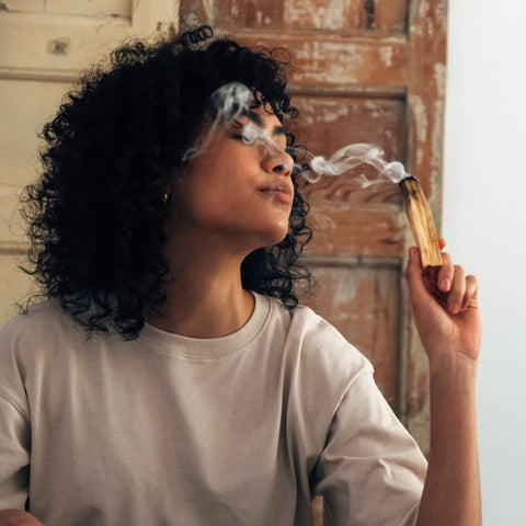 A woman with curly black hair wearing a light beige t-shirt holds a burning sage smudge stick near her face, with smoke drifting across her face and hair, set against a rustic wooden door and white wall background.