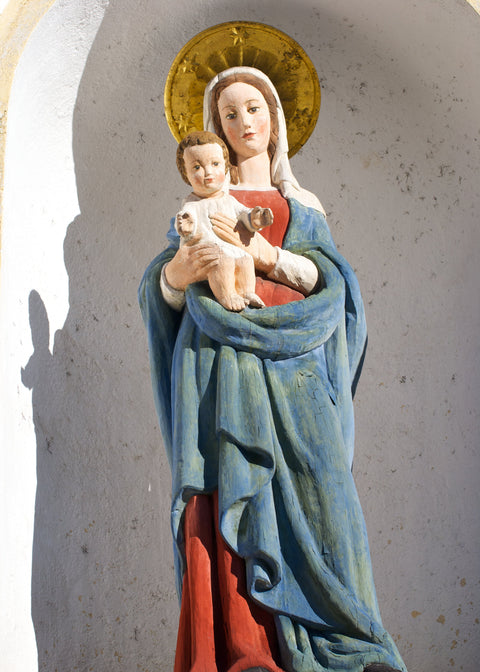 A painted religious statue of the Virgin Mary holding the infant Jesus, shown against a textured light gray wall. Mary wears a blue cloak over a red dress, with a gold halo behind her head.