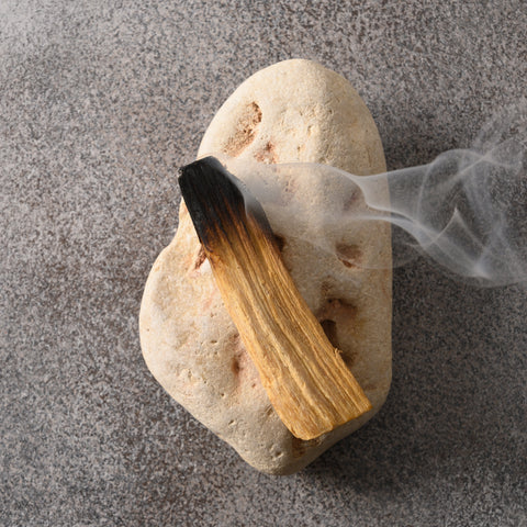 A heart-shaped beige stone with a smudging bundle of dried grass and blackened tip resting on it, with wisps of white smoke curling to the right, set against a textured gray background.