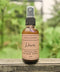 A 2 oz amber glass spray bottle of Soulgasm Weaver Smokeless Sacred Spray, resting on a weathered wooden surface with a soft-focus green garden background.