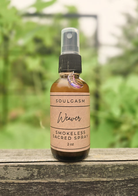 A 2 oz amber glass spray bottle of Soulgasm Weaver Smokeless Sacred Spray, resting on a weathered wooden surface with a soft-focus green garden background.
